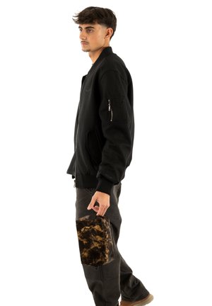 Črna bomber jakna z rebrastimi manšetami in žepom z zadrgo, kombinirana s temno sivimi hlačami. Model drži rjavo marmorirano torbico.