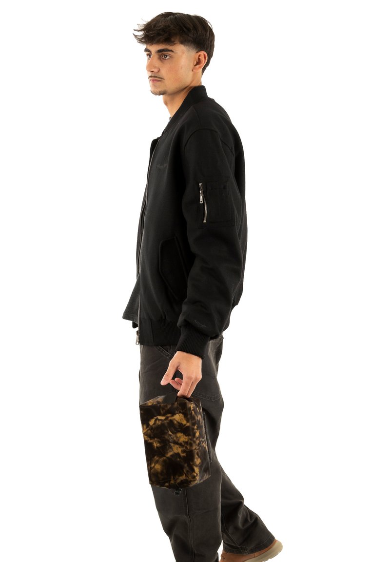 Giacca bomber nera con polsini a costine e tasca con zip, abbinata a pantaloni grigio scuro. Il modello tiene in mano una pochette marrone marmorizzata.