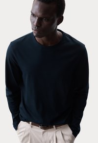 Langarmshirt in Marineblau aus weichem Stoff mit Rundhalsausschnitt, kombiniert mit hellbeigen Hosen und einem braunen Gürtel. Keine sichtbaren Muster oder Akzente.