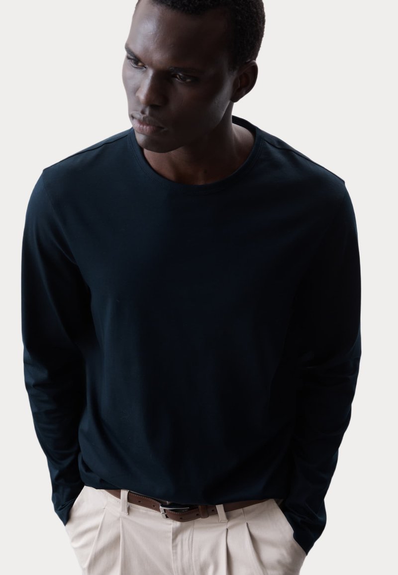 Langarmshirt in Marineblau aus weichem Stoff mit Rundhalsausschnitt, kombiniert mit hellbeigen Hosen und einem braunen Gürtel. Keine sichtbaren Muster oder Akzente.