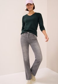 Tmavě zelený dlouhý rukáv s výstřihem do V, kombinovaný se světle šedými rozšířenými džíny. Modelka má na sobě růžovou čepici a béžové tenisky.