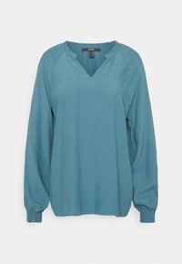 Blouse à manches longues en V, couleur bleu-vert, avec une finition texturée, des poignets froncés et un léger détail de volant au niveau du col. Tissu doux et léger.