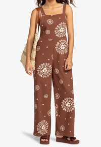 Bruine jumpsuit met grote witte bloemmotieven, vierkante halslijn, wijde pijpen en verstelbare bandjes, gecombineerd met bruine platform sandalen en een rieten tas.