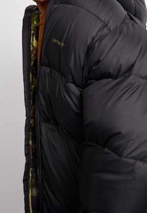 Gros plan d'une personne portant une doudoune noire avec une doublure intérieure camouflage et un pull marron partiellement visible.