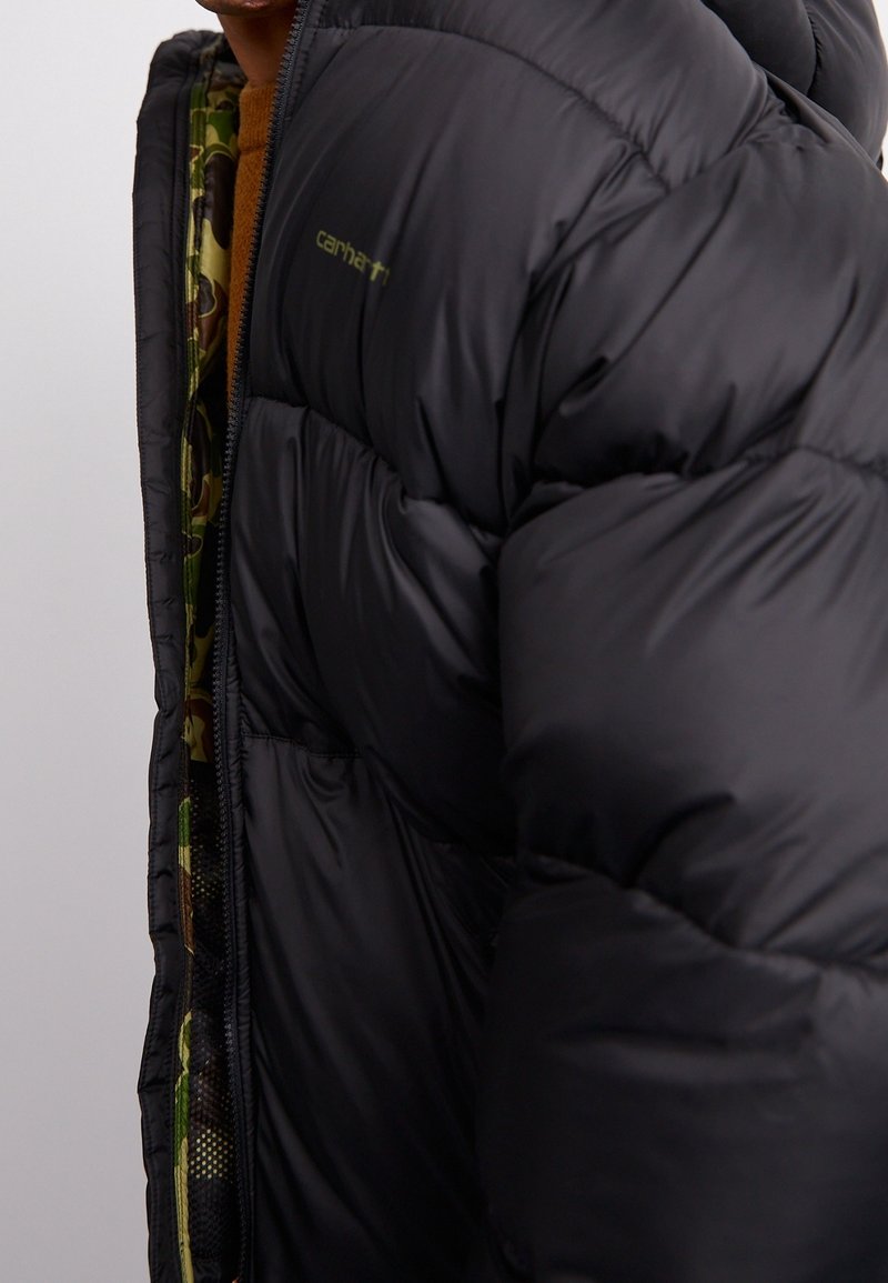 Gros plan d'une personne portant une doudoune noire avec une doublure intérieure camouflage et un pull marron partiellement visible.