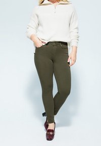 Jeans skinny verde oliva con una textura suave, combinados con un suéter de punto blanco con cremallera y zapatos bajos burdeos.