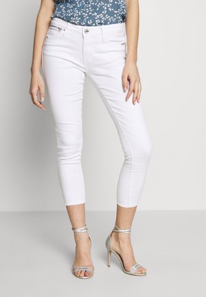 Une personne portant un pantalon skinny court blanc, un haut bleu à motifs floraux et des sandales argentées à talons hauts, se tenant sur un fond neutre.