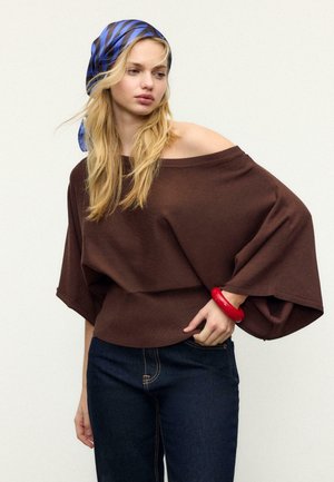 Plavolasa ženska z modro-črnim črtastim ruto na glavi nosi rjav vrh brez ramen, temne kavbojke in rdečo zapestnico, pozira z eno roko na boku.