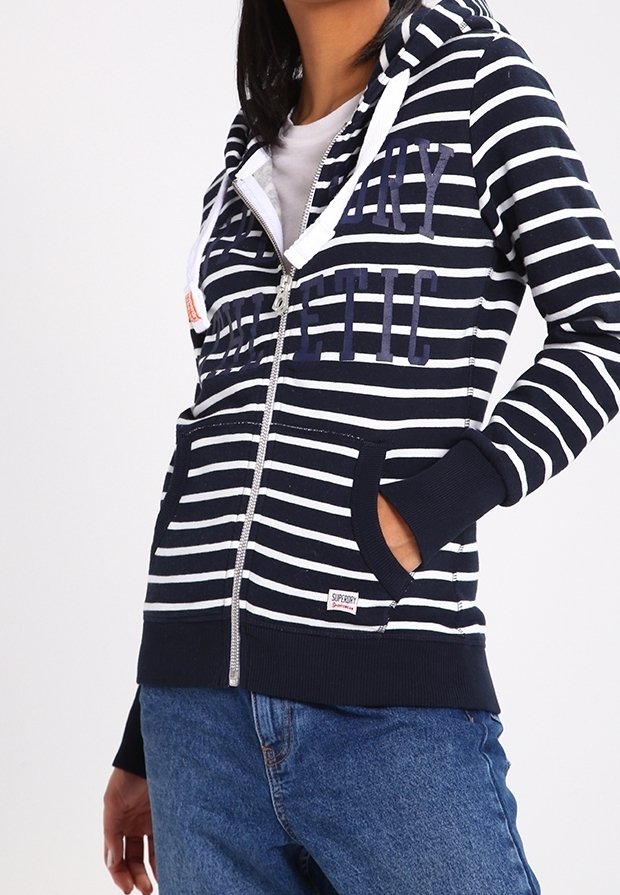 Personne portant un sweat à capuche zippé rayé bleu marine et blanc avec des poches et un jean bleu, debout avec une main dans la poche sur un fond uni.