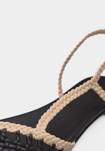 Μαύρο σανδάλι με σόλα, με μπεζ πλεγμένο λουράκι και λουρί στον αστράγαλο, εμφανίζεται σε κοντινό πλάνο σε λευκό φόντο.