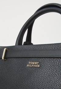 Sort textureret lædertaske med et guld-tone "TOMMY HILFIGER" logo, runde sorte håndtag og et slankt, struktureret design.