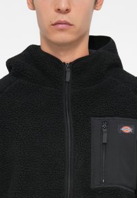 Veste en polaire noire avec un col haut, une fermeture éclair à l'avant et une poche zippée sur la poitrine avec un patch logo.