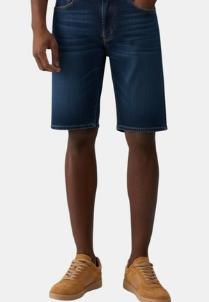 Uomo con pantaloncini di jeans blu scuro e sneakers in camoscio beige, in piedi con le mani in tasca.