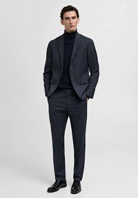 Dark gray suit with a textured pattern, single-breasted jacket, slim-fit trousers, and a black turtleneck underneath. Black leather shoes.