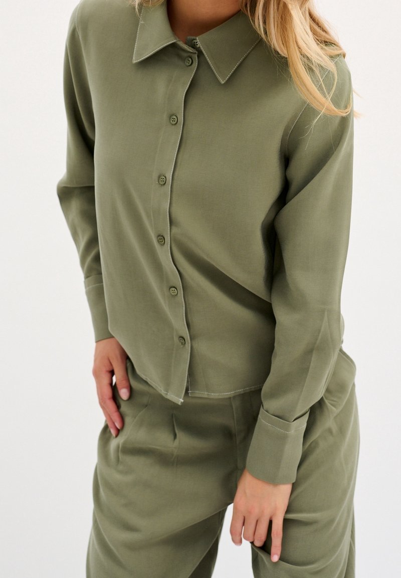 Femme portant une chemise boutonnée vert olive avec col et un pantalon assorti, une main posée sur la hanche et l'autre le long du corps.