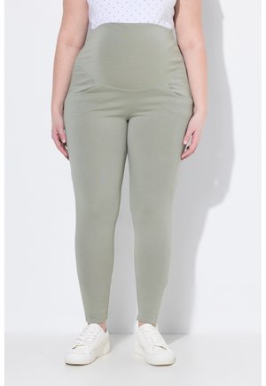 Kobieta ubrana w jasnozielone legginsy ciążowe z wysokim stanem, zestawione z białymi trampkami i białą bluzką w groszki na białym tle.