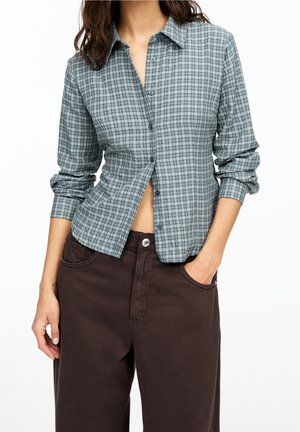 Chemise à carreaux bleue et verte, style boutonné, manches longues, tissu doux, coupe décontractée ; portée avec un pantalon marron foncé.