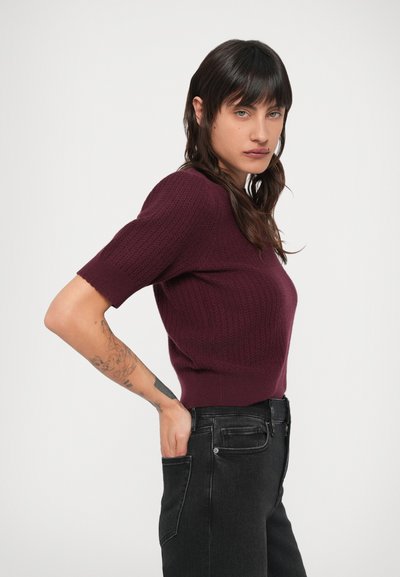 Bordó kötött felső rövid ujjakkal és testhezálló szabással, magas derekú fekete farmerral párosítva. Texturált anyag bordázott mintával.