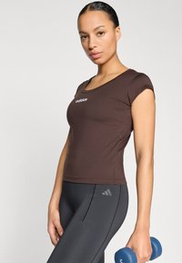 Brown athletic t-shirt with a scoop neckline and short sleeves, featuring a white logo. Model holds a blue dumbbell, paired with black leggings.