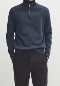 Pull-over en maille bleu marine avec un col haut et une fermeture éclair. Poignets et ourlet côtelés. Associé à un pantalon sombre texturé, mains dans les poches.