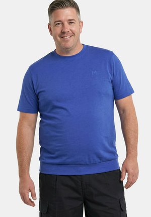 Lachende man die een blauw T-shirt met korte mouwen en een ronde hals en zwarte cargoshorts draagt, tegen een effen lichte achtergrond.