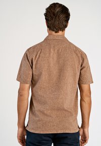 Kurzärmeliges, braunes Hemd mit Kragen, strukturiertem Stoff, sichtbaren Nahtdetails und seitlichen Schlitzen am Saum für bessere Bewegungsfreiheit.