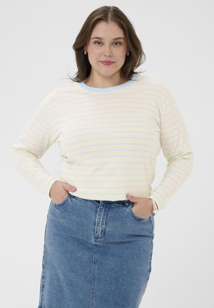 Femme aux cheveux châtains moyens portant une chemise à manches longues rayée jaune pastel et bleue, jupe en denim bleu, mains dans les poches.