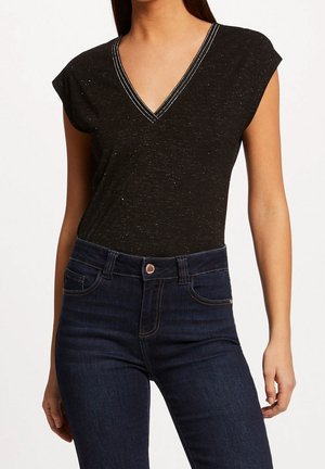 Femme portant un haut sans manches noir à pois avec un col en V, rentré dans un jean taille haute bleu foncé avec poches avant et fermeture par bouton.