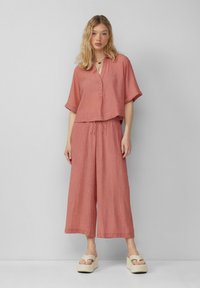Roodbruin, kort geknipt overhemd met knopen en korte mouwen, gecombineerd met bijpassende wijde broek. Zacht, textuurstof; minimalistisch ontwerp.