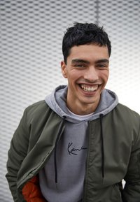 Olivgrüne Bomberjacke über einem grauen Hoodie mit einem schwarzen Signaturdruck. Der strukturierte Hintergrund ist hellgrau mit einer gemusterten Oberfläche.