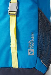 Sininen Jack Wolfskin -reppu, jossa keltainen hihna ja harmaa muovisolki sekä näkyvä musta verkkotasku sivulla.