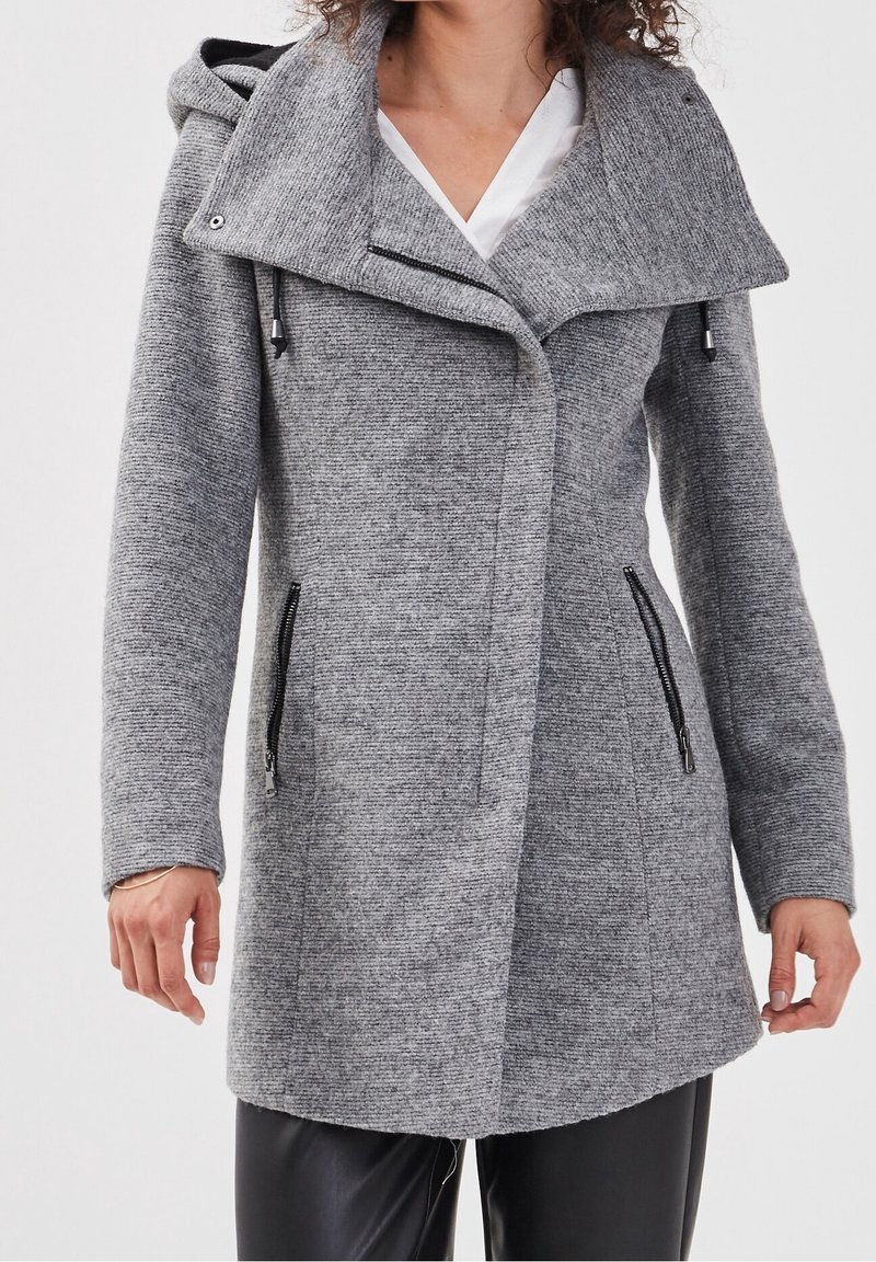 Manteau gris texturé avec un grand col, des fermetures éclair à l'avant et une capuche. Comprend des poches latérales avec des fermetures éclair noires et une coupe cintrée.