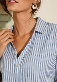 Camicia a righe blu e bianche con scollo a V e piccolo bottone bianco. Presenta un colletto e un tessuto morbido, abbinata a orecchini a cerchio.