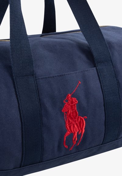 Marineblå canvas duffelbag med rød brodert polo-spillerlogo, kontrasterende håndtak og glidelås. Strukturert overflate og forsterkede sømmer.