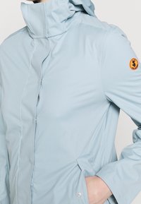 Personne portant une veste à capuche bleu clair avec la main dans la poche latérale et un petit écusson rond orange sur la manche supérieure.