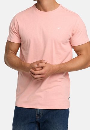 Homme portant un t-shirt rose clair à manches courtes avec un petit logo blanc sur la poitrine, les mains jointes, associé à un jean bleu.