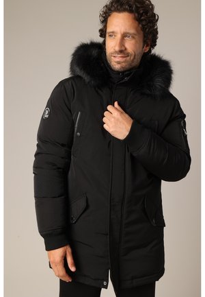 Man draagt een zwarte gewatteerde winterjas met een bontkraag, staand met één hand bij de borst tegen een neutrale achtergrond.