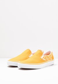 Baskets jaunes à enfiler en toile. Semelle en caoutchouc blanche. Un côté présente un motif de logo répété en orange sur fond blanc.