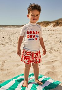Vit kortärmad rashguard med röd och blå text, tillsammans med röda simshorts med mönster i abstrakt design. Står på en handduk med gröna ränder.