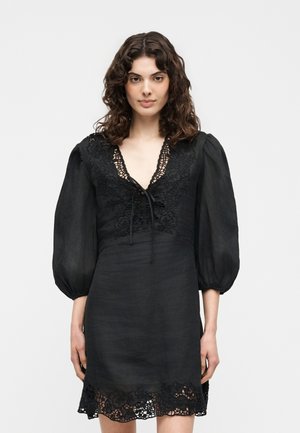 Γυναίκα με κατσαρά μαλλιά που φοράει μαύρο φόρεμα με δαντελένιες λεπτομέρειες, φουσκωτά μανίκια μέχρι τον αγκώνα και V-λαιμόκοψη με κορδόνια.