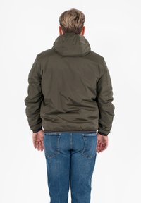Giacca imbottita verde oliva con cappuccio, polsini e orlo elasticizzati. Indossata sopra dei jeans blu, vista da dietro, mettendo in evidenza il colletto e le cuciture.