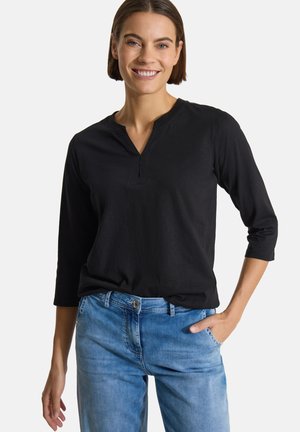 Femme souriante portant un haut noir à manches trois-quarts et un jean bleu, une main dans la poche, sur un fond uni.