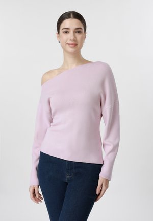 Γυναίκα με ανοιχτό ροζ πουλόβερ χωρίς ώμους και σκούρο μπλε τζιν, που στέκεται μπροστά σε απλό φωτεινό φόντο, χαμογελώντας απαλά.