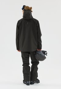 Schwarze wasserdichte Skijacke mit Kapuze, kombiniert mit passenden Hosen. Die Person hält einen grauen Helm und trägt schwarze Skischuhe.