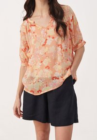 Blouse à motifs floraux dans des teintes de pêche doux et de pastel, en tissu léger, avec un décolleté en V et des manches courtes retroussées. Associée à un short foncé.
