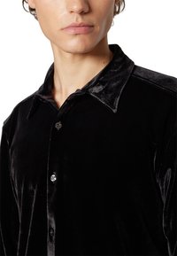 Jeune homme portant une chemise en velours noire boutonnée avec un col, visible du cou jusqu'au milieu du torse sur fond blanc.