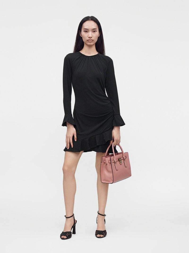 Jeune femme debout, portant une robe noire à manches longues avec des volants, des sandales à talons noirs, tenant un sac à main rose dans la main droite.