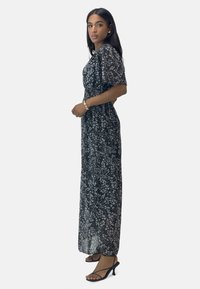 Langes schwarzes Blumenkleid aus leichtem, halbtransparentem Stoff, mit Puffärmeln, tailliertem Bund und schwingendem Schnitt. Getragen mit schwarzen Sandalen.