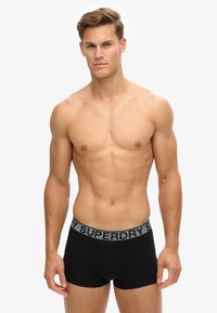 Bóxer negro de corte trunk con una cinturilla gris que presenta el logo "SUPERDRY". Tejido suave con un ajuste ceñido y un diseño minimalista.