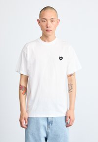 White cotton t-shirt with short sleeves, round neck, and a small black heart logo on the left chest. Model wears light blue denim bottoms.